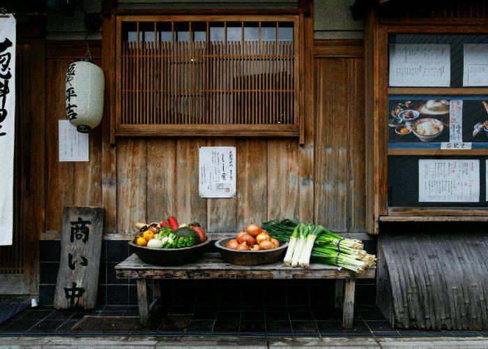 Fresh vegetables for sale on a bench outside a traditional Japanese restaurant, complete with a paper lantern.